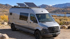 Mercedes Sprinter camper parked beside a lake with two roof solar panels tilted toward the sun