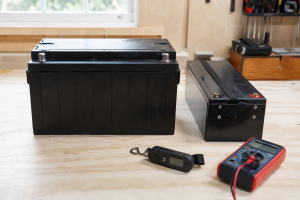 A large and a slim 12V battery side by side on a workbench, next to a luggage scale and a multimeter