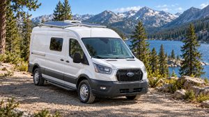 White Ford Transit camper van with two roof-mounted solar panels at a mountain lake campsite