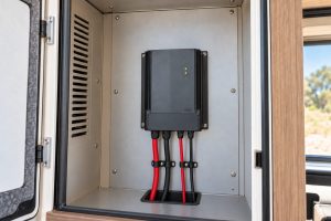 Solar charge controller standing upright on a metal backing plate in a vented cabinet, with its four cables clamped and routed down through a rubber-lined floor opening