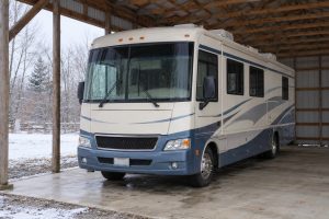 Cream and blue Class A motorhome parked under a timber shelter, with snow on the ground outside