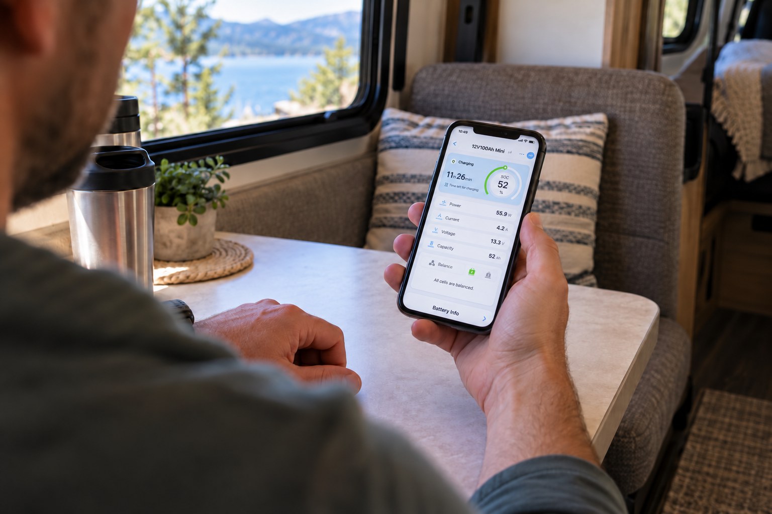 Traveler checking a detailed battery app on a phone at an RV dinette, with charge status, voltage, current and remaining capacity on the screen