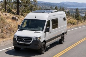 White Mercedes-Benz Sprinter camper van with low-profile roof solar panels driving on a sunlit country road